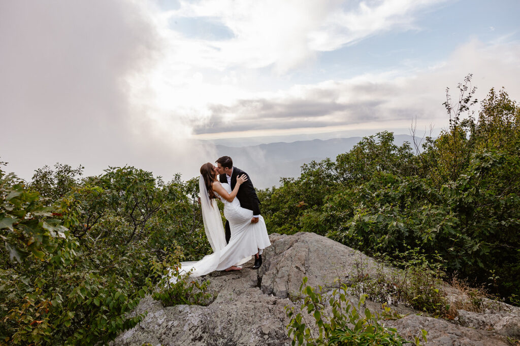 The bride and groom share a romantic kiss on top of a rocky hill with the misty mountains and clouds in the background. The bride's long veil flows behind her as they stand surrounded by lush greenery.