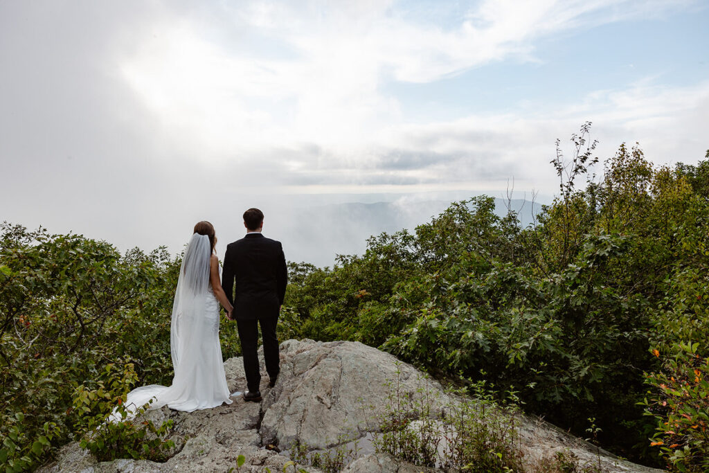 The bride and groom stand together on a rocky ledge, hand in hand, looking out over the misty mountains in the distance. Their backs are to the camera, with the bride's veil trailing behind her.