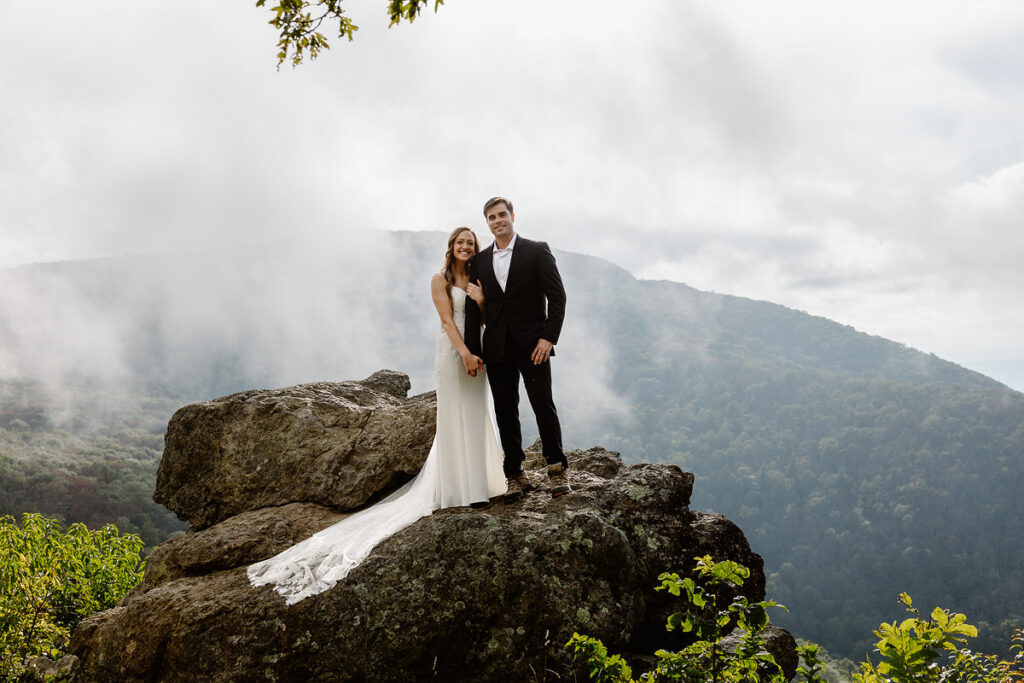 The bride and groom stand on a large rock, with the bride’s lace train flowing down the side. They smile warmly at the camera with foggy mountains in the background, creating a serene and romantic atmosphere.