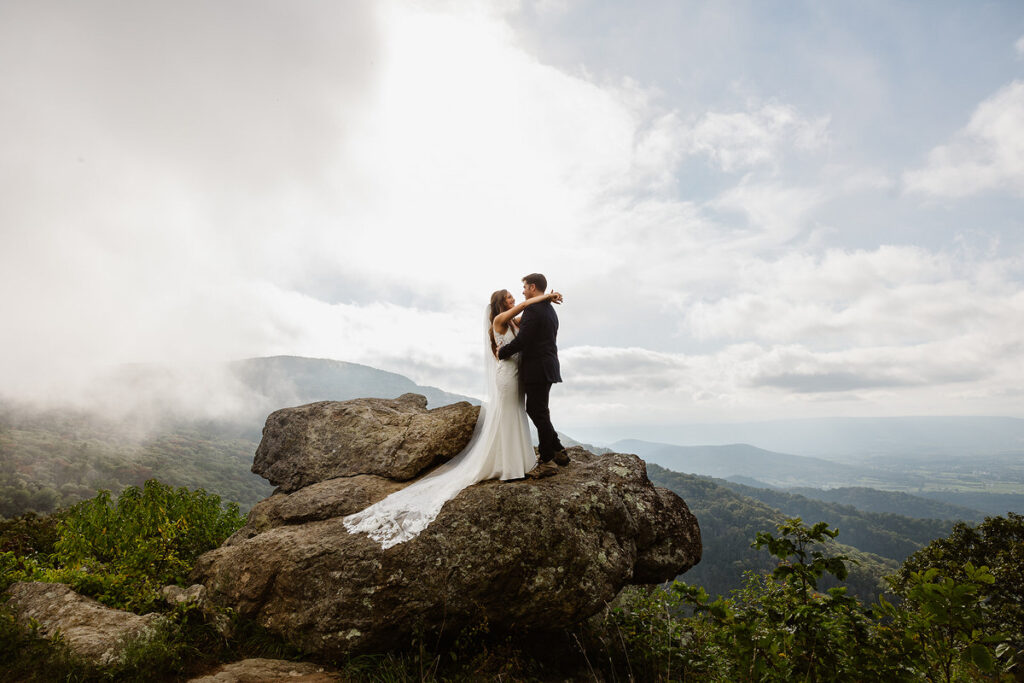 The bride and groom stand together on a large rock, embracing each other with a vast mountainous landscape and clouds in the background. The bride’s long white dress trails behind her as they share a romantic moment with the scenic Shenandoah National Park view.