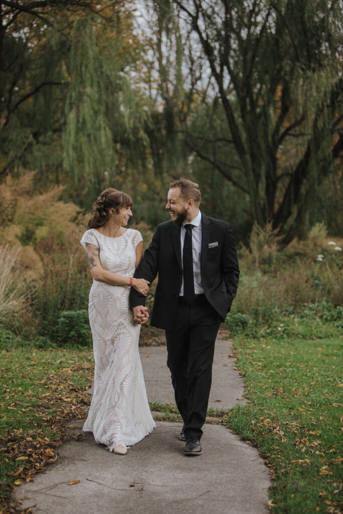 A bride and groom walk hand-in-hand along a garden path surrounded by lush greenery. The bride wears an elegant lace gown with cap sleeves, while the groom looks smart in a black suit and tie. They gaze at each other with smiles, suggesting a moment of shared happiness and connection.