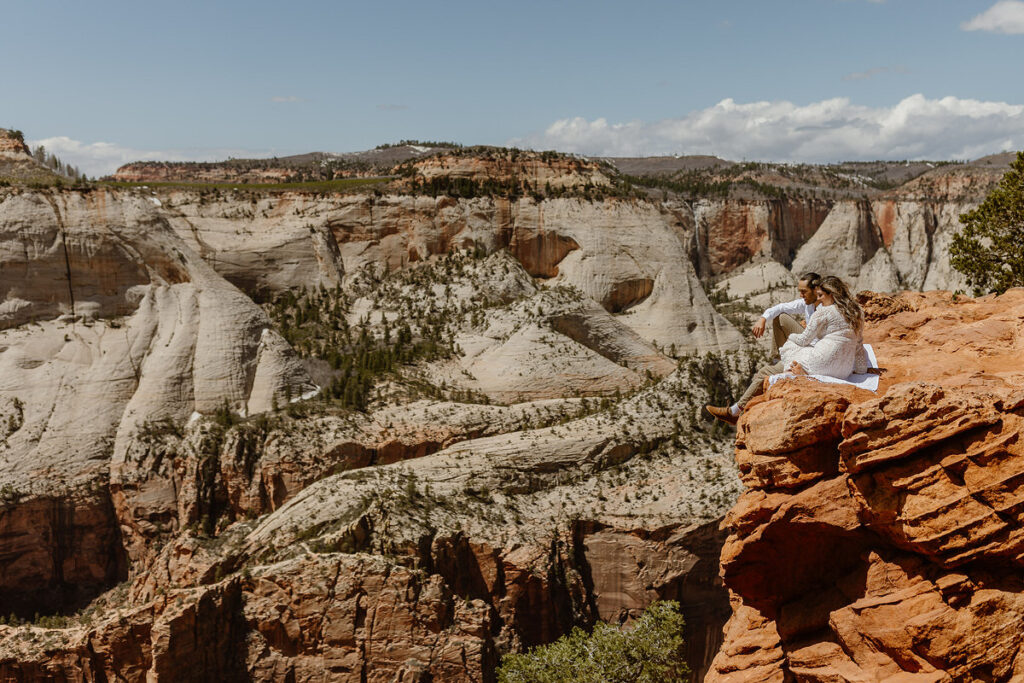 A couple sits on the edge of a cliff overlooking a stunning canyon landscape, possibly during an elopement or intimate wedding. The bride wears a lace dress, and they seem to be enjoying the breathtaking view of the expansive, rugged rock formations and clear sky. The setting suggests adventure and a love for nature.