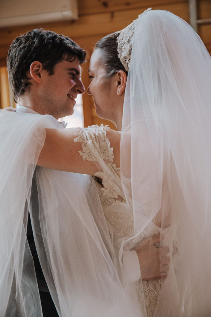 A close-up, intimate shot of a bride and groom sharing a tender moment indoors. The bride wears a detailed, lace veil that wraps around her shoulders, and the groom is in a suit. Their faces are close together, and they both appear to be smiling, capturing the joy and love of the occasion.