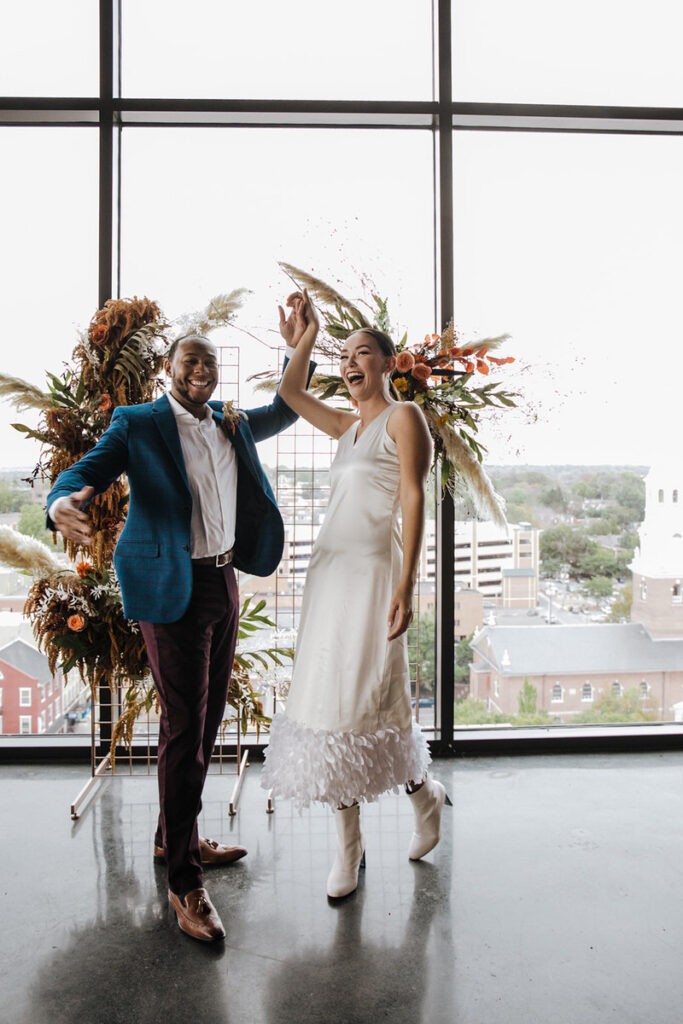 A couple dressed in wedding attire shares a joyful moment, possibly in an urban indoor space. The groom wears a stylish blue blazer while the bride is in a simple white dress with white ankle boots. They seem to be dancing or celebrating, with a cityscape visible through large windows behind them, and decorative floral arrangements in the background.