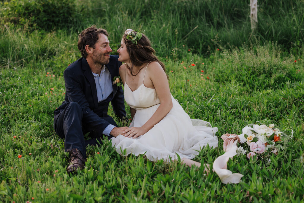 A couple sitting together on a grassy field. The bride is wearing a flowing white dress and a floral crown, while the groom is dressed in a dark suit. They look lovingly at each other, surrounded by lush greenery and a bouquet of flowers lying nearby.