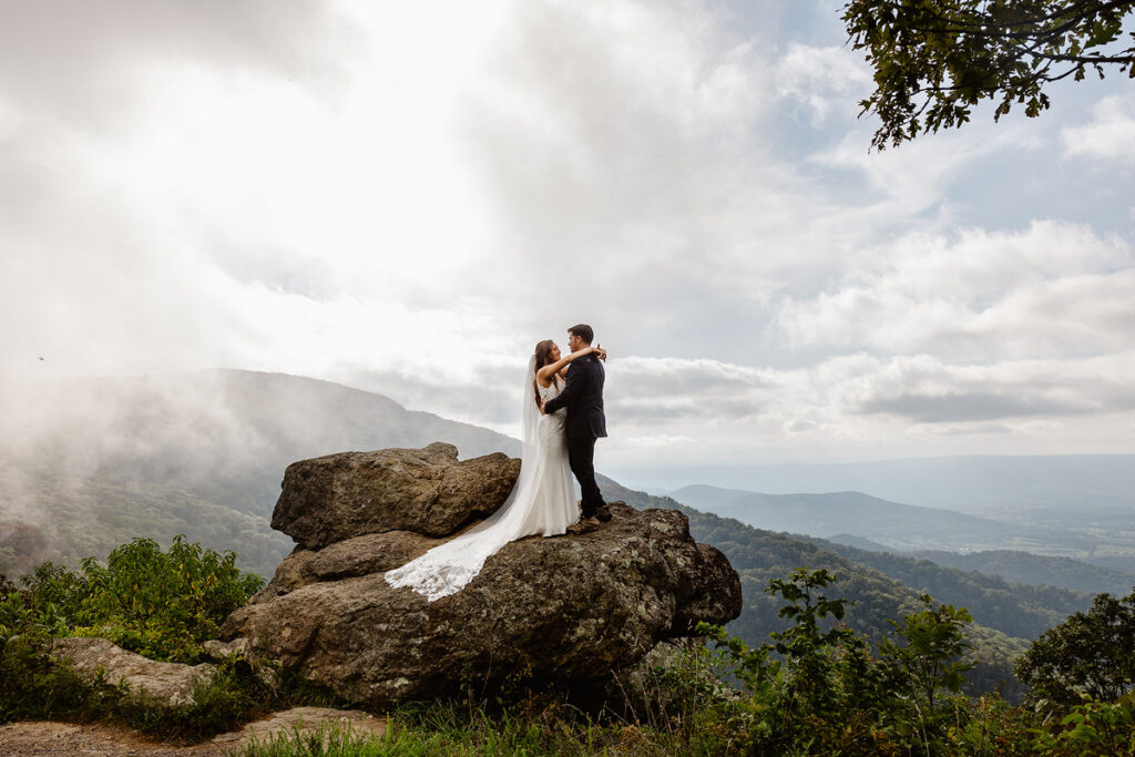 A bride and groom stand on a large rock with a misty mountain landscape in the background. The bride’s long dress flows behind her, and they embrace as they take in the expansive view. This scene captures a moment of romantic connection in a dramatic, natural setting, emphasizing the beauty of an adventurous elopement or intimate wedding.