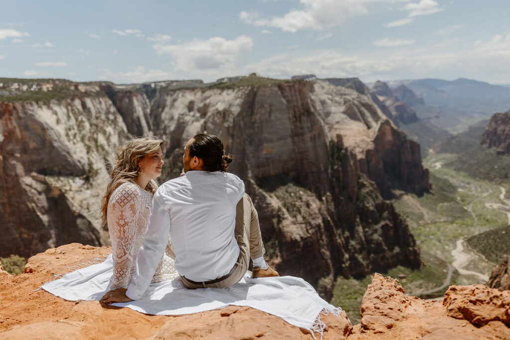 A couple sits on a blanket at the edge of a canyon, looking into each other’s eyes. The bride wears a long-sleeved lace dress, and the groom is in a white shirt with khaki pants. The expansive canyon and bright sky in the background make the setting feel adventurous and romantic.