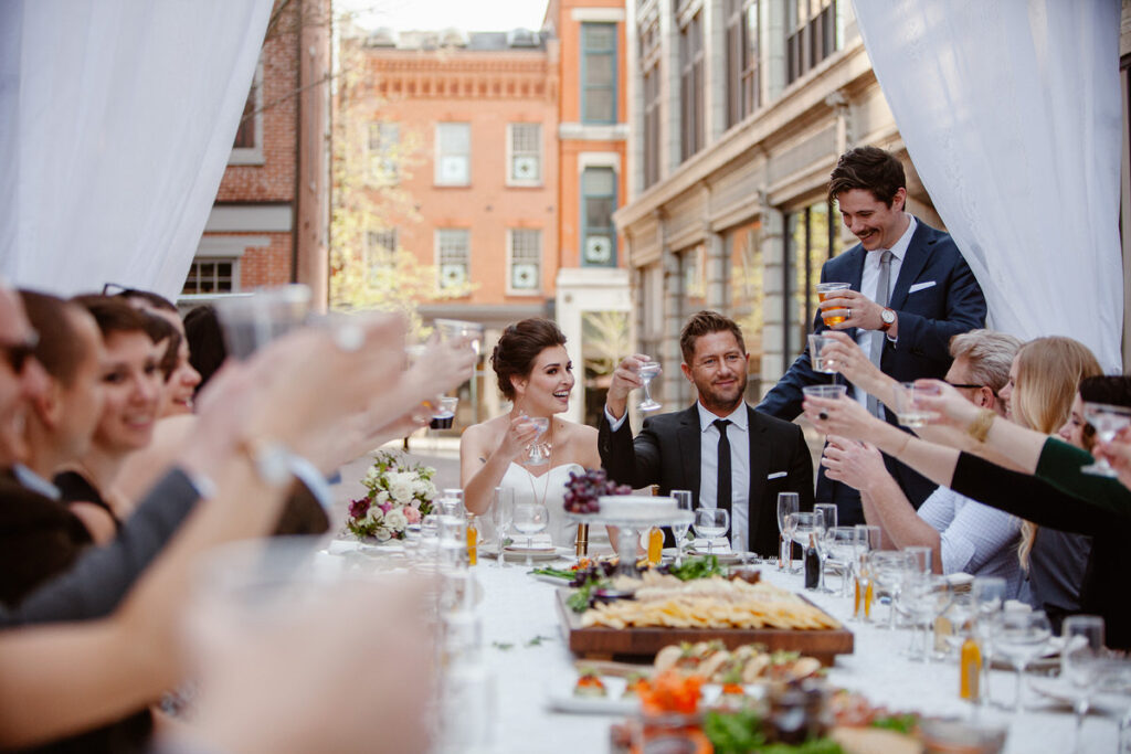 A joyful wedding celebration takes place at an outdoor reception with a long dining table surrounded by guests raising glasses in a toast. The bride and groom, both dressed elegantly, are seated at the head of the table, with the groom standing to make a speech. The urban backdrop and relaxed, festive atmosphere indicate a small, intimate wedding gathering.