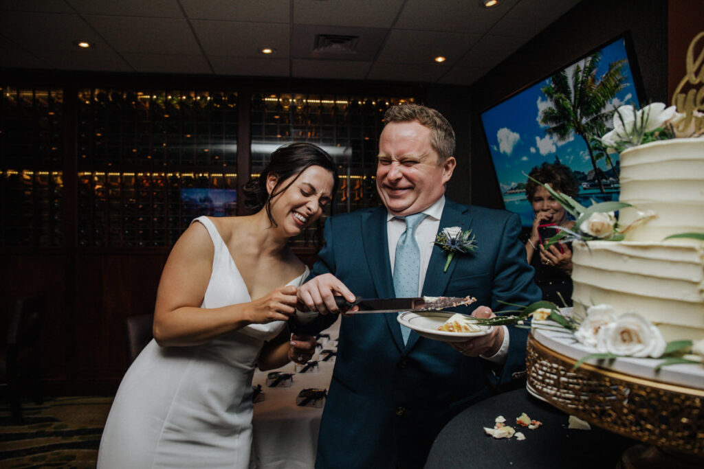 A bride and groom share a playful moment cutting their wedding cake indoors. The bride, in a white dress, and the groom, in a dark suit, both laugh as they hold the cake knife together. The scene captures a lighthearted and fun element of their celebration, with a multi-tiered cake in the foreground.