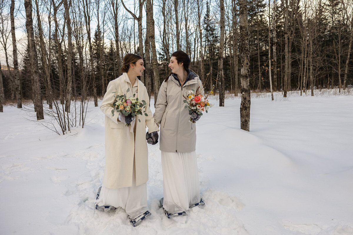 Two brides dressed in white wedding gowns walk hand-in-hand through a snowy forest, both wearing warm coats. They hold bouquets of flowers with colorful blooms and smile at each other. Their expressions and attire reflect a winter elopement or wedding, conveying a sense of warmth and love despite the cold surroundings.