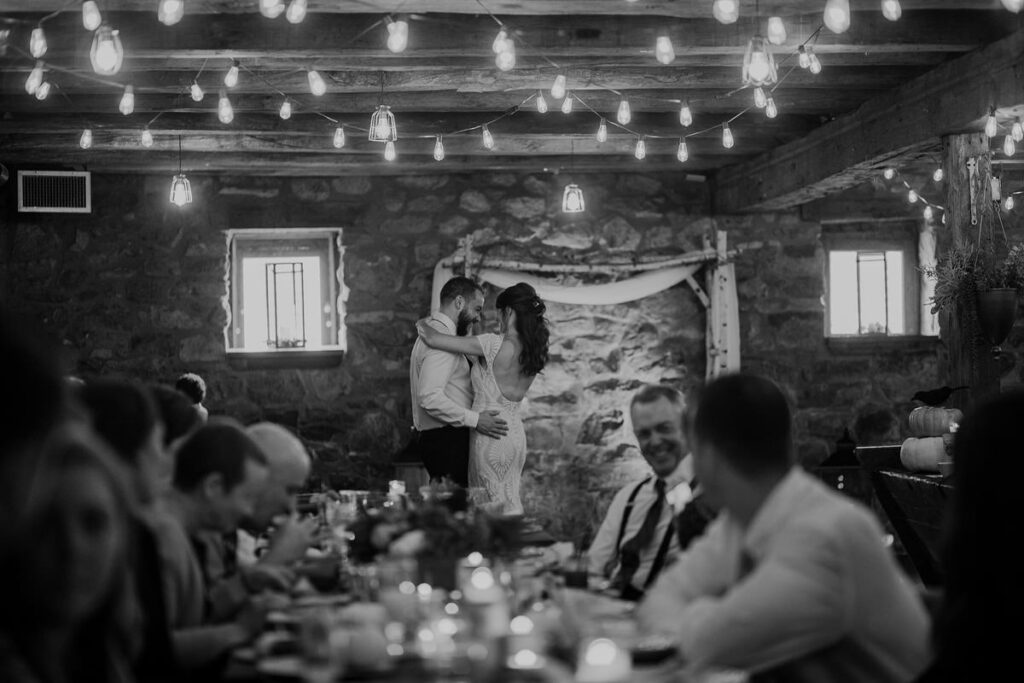 A black and white photo of a bride and groom embracing with guests sitting at a table out of focus in the forefront