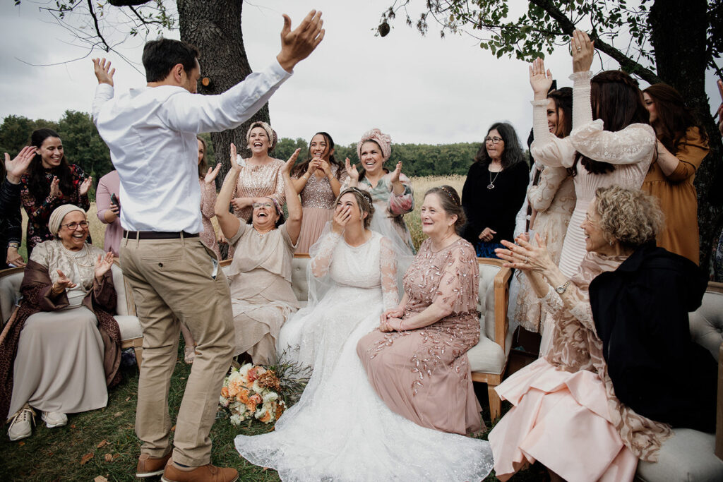 A bride and her bridesmaids laugh as the groom entertains them in an outdoor setting