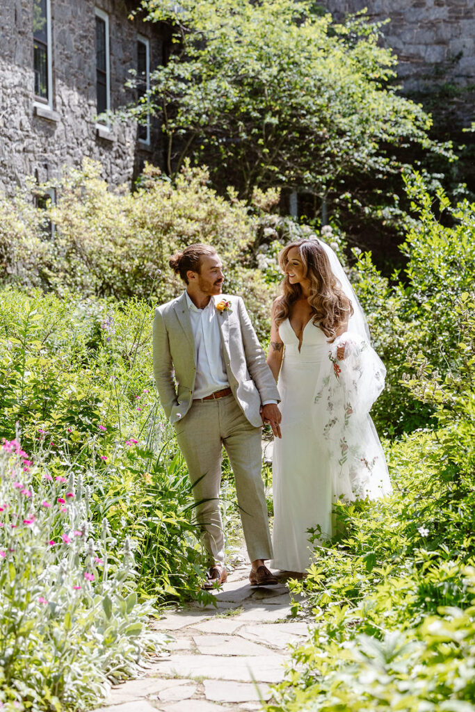 A bride and groom in a tan suit walk through a wildflower garden holding hands