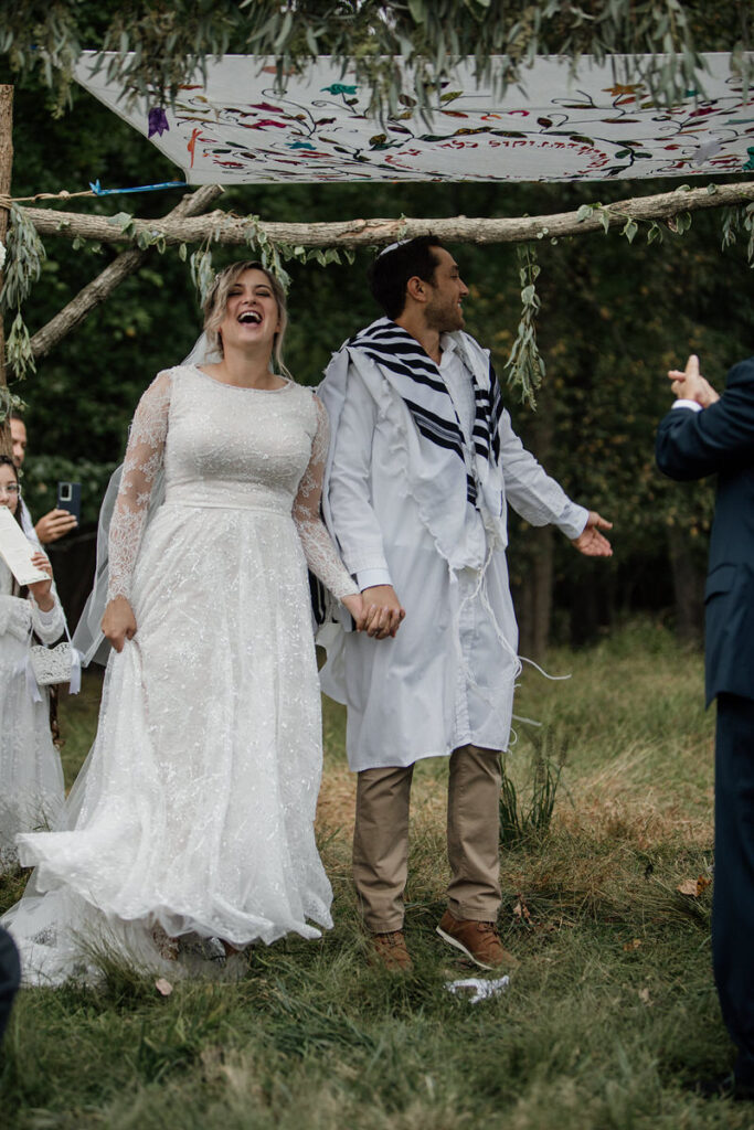 A bride and groom stand under a canopy made of tree branches and fabric holding hands and laughing