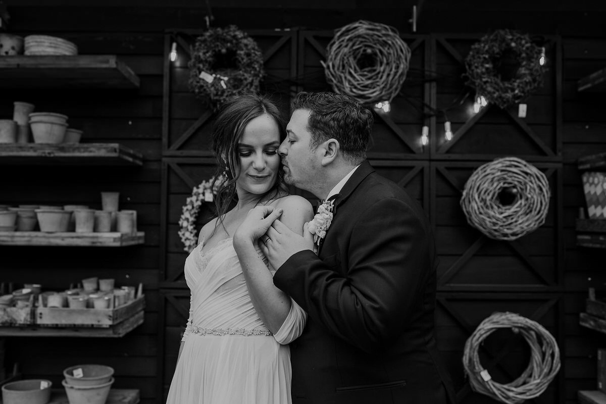 A black and white photo of a groom gently kissing the bride on the shoulder, in an intimate moment surrounded by shelves of pots and hanging wreaths. The bride is dressed in an off-shoulder gown, and the groom wears a suit with a boutonniere.