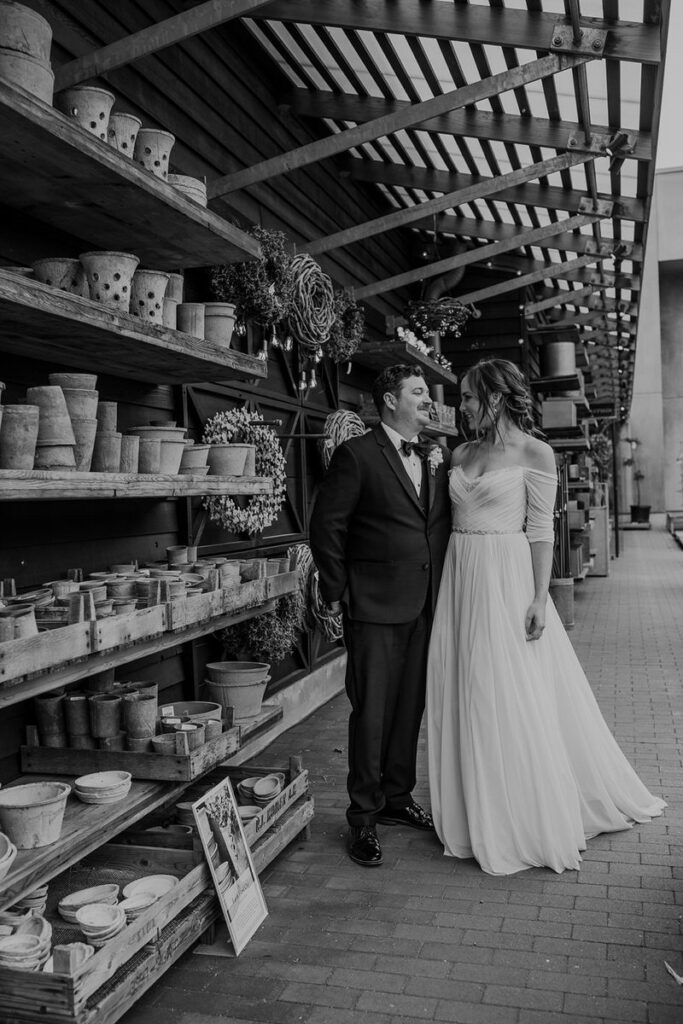 A black and white photo showing a couple standing close together in a rustic setting filled with rows of pots and wreaths on shelves. The bride is in an elegant dress, and the groom is in a formal suit, gazing into each other's eyes.