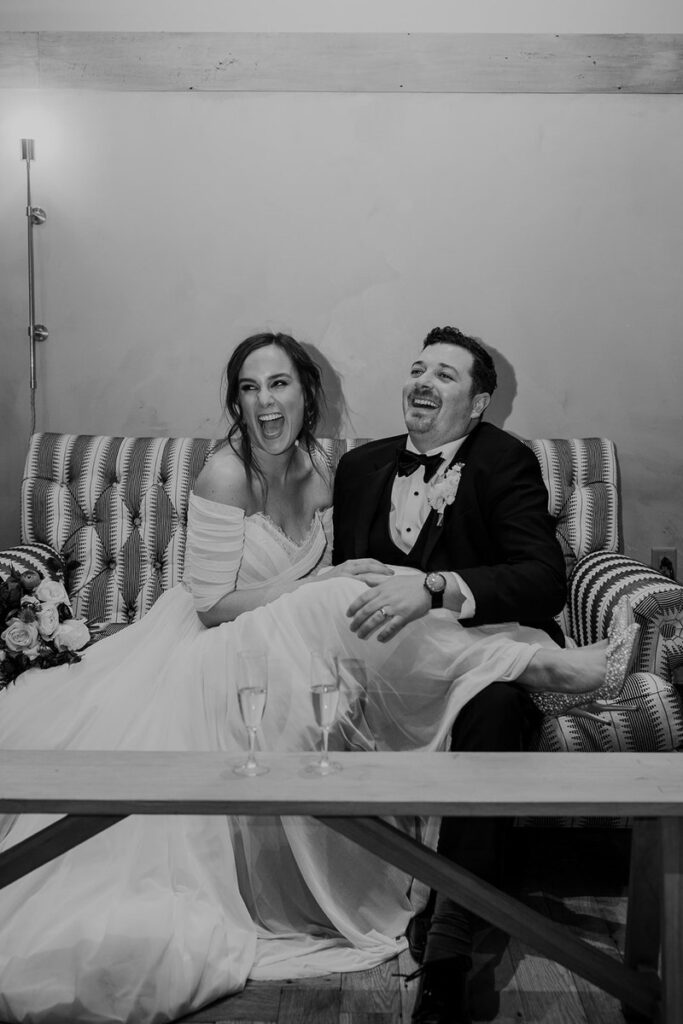 A black and white photo of a bride and groom sitting on a striped couch laughing