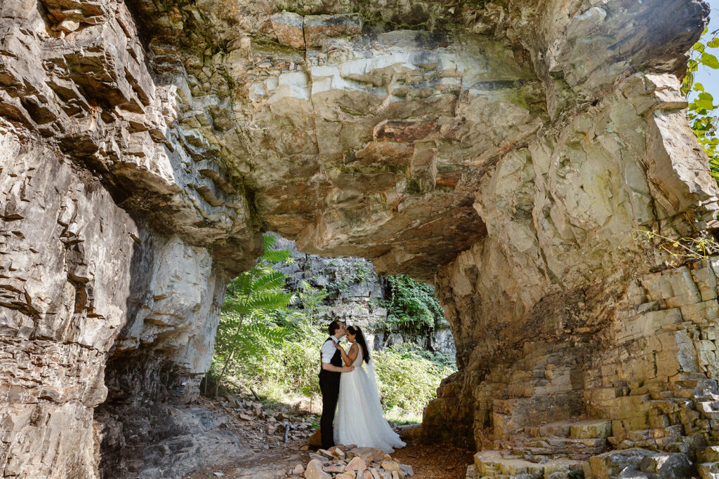 A couple stands in a cave at Natural Chimneys Park. They are a bride and groom, and they just got married. She is wearing a tiara and the groom has on a bowtie and a wool suit. They are standing facing each other, and the groom is kissing the forehead of the bride.