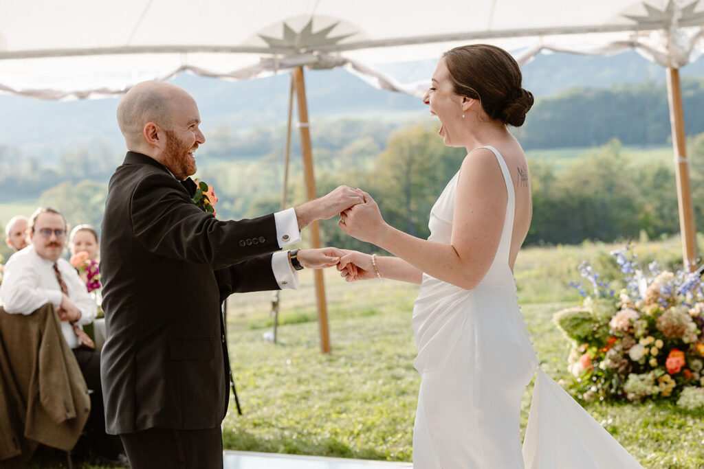 A bride and groom during their first dance. They are dancing under a sail cloth tent. They are both smiling with excitement. In the background you can see trees and mountains. The bride is wearing a white satin dress, and the groom is wearing a black tux