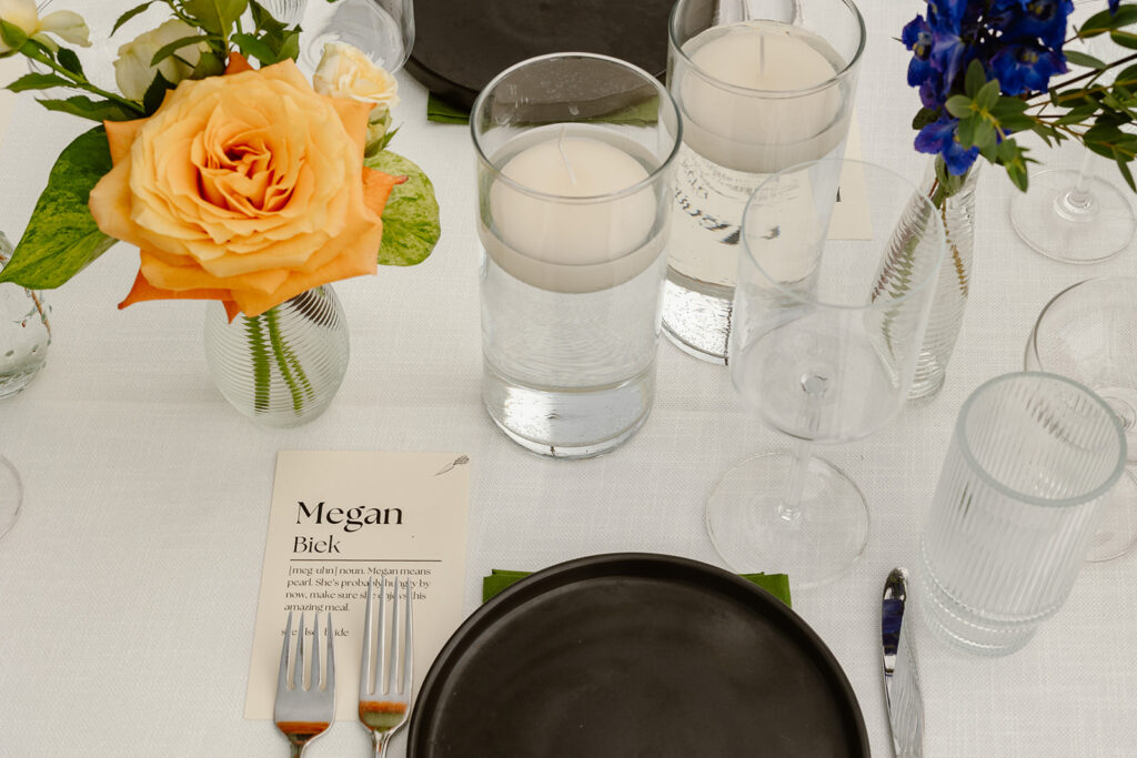 A photo of some of the details of the table setting at a reception. There is a large peach roase in a vase along with another vase filled with wildflowers. There is also a modern black plate, and a name card