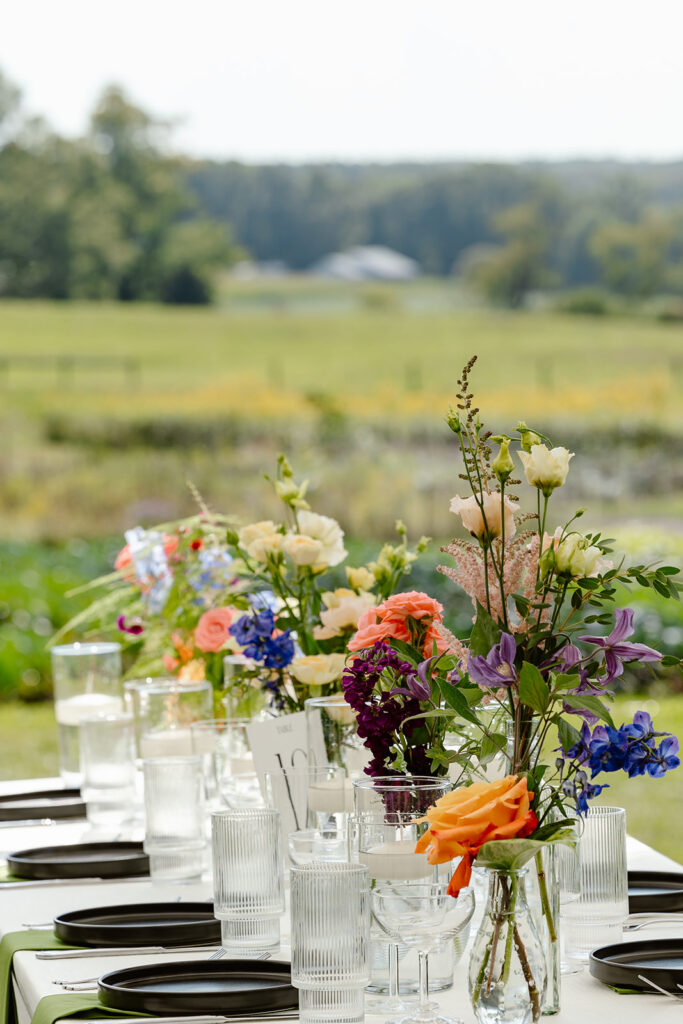 A photo of the tables at a reception. Their s a mix of roses and  colorful wildflowers, and in the background you can see farmland