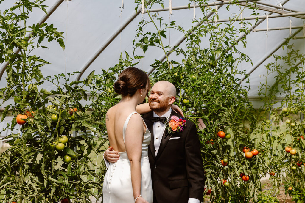 A bride and groom embrace after seeing each for the first time on their wedding day. They are in a tomato greenhouse. The bride is wearing a sleeveless satin dress, and the groom is wearing a black tux