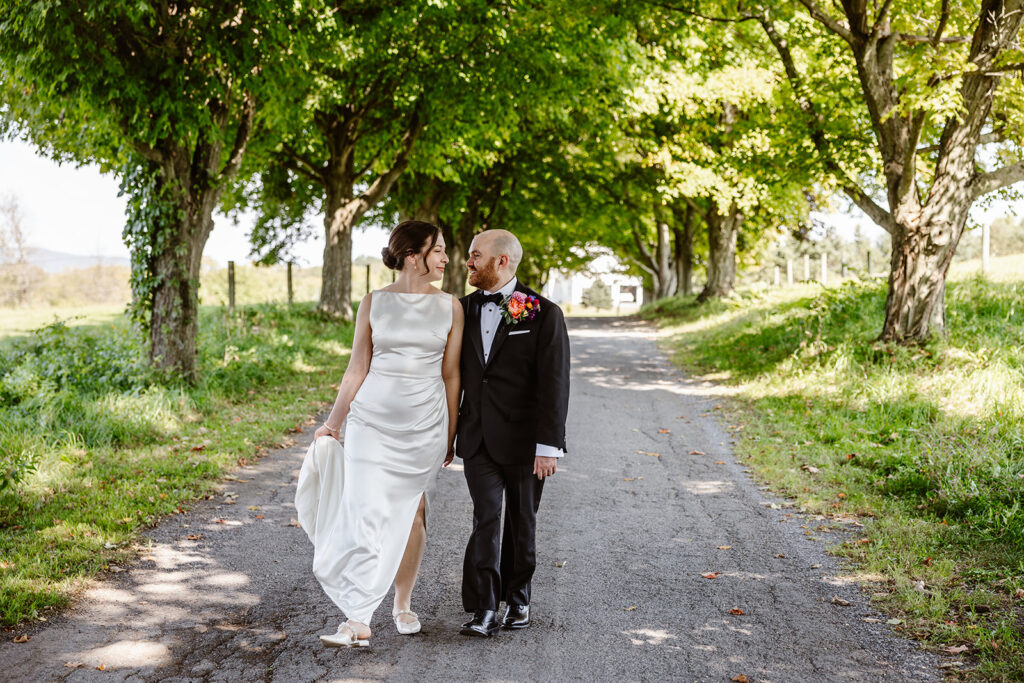 A bride and groom walking hand in hand on a driveway. There are trees on both sides. The bride and groom are looking at each other lovingly. She bride is wearing a sleeveless satin dress, and the groom is wearing a black tux