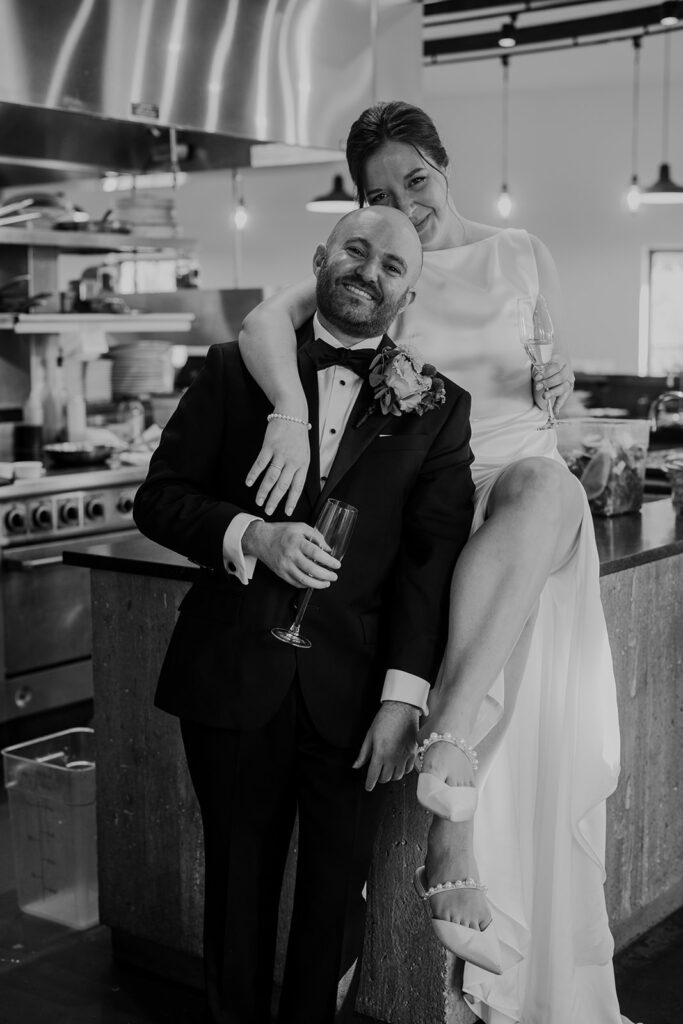 A black and white photo of a bride and groom sitting in a prep kitchen. The bride is sitting on the countertop with her legs crossed, and the groom is standing next her leaning in. Both of them are holding wine glasses, and the bride has her arm around the groom.