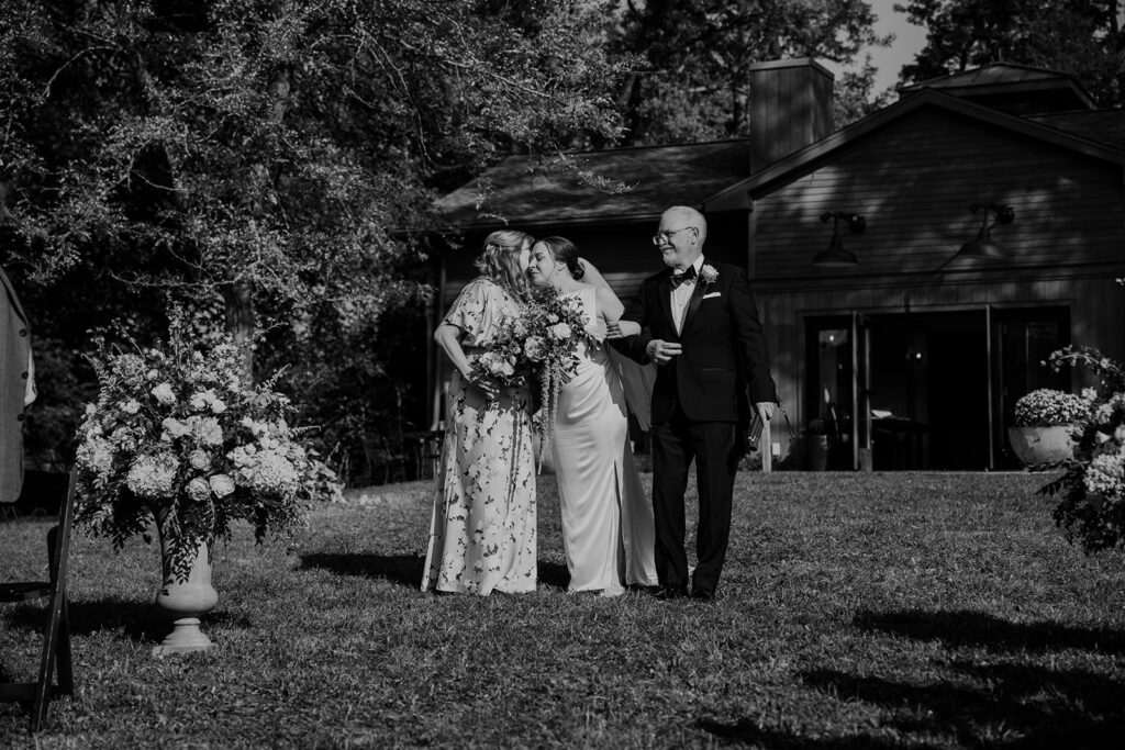 A black and white photo of a bride and her parents walking down the grass aisle at the intimate outdoor ceremony. The Brides mom is kissing the bride on the cheek. There are large flower arrangements on each side of the aisle.