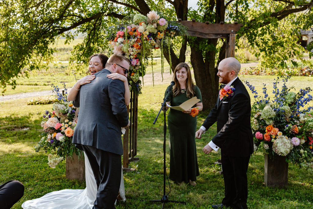 The bride hugs a man who just found out that he is the ring bearer for the wedding. The groom and officiant are looking at them smiling.