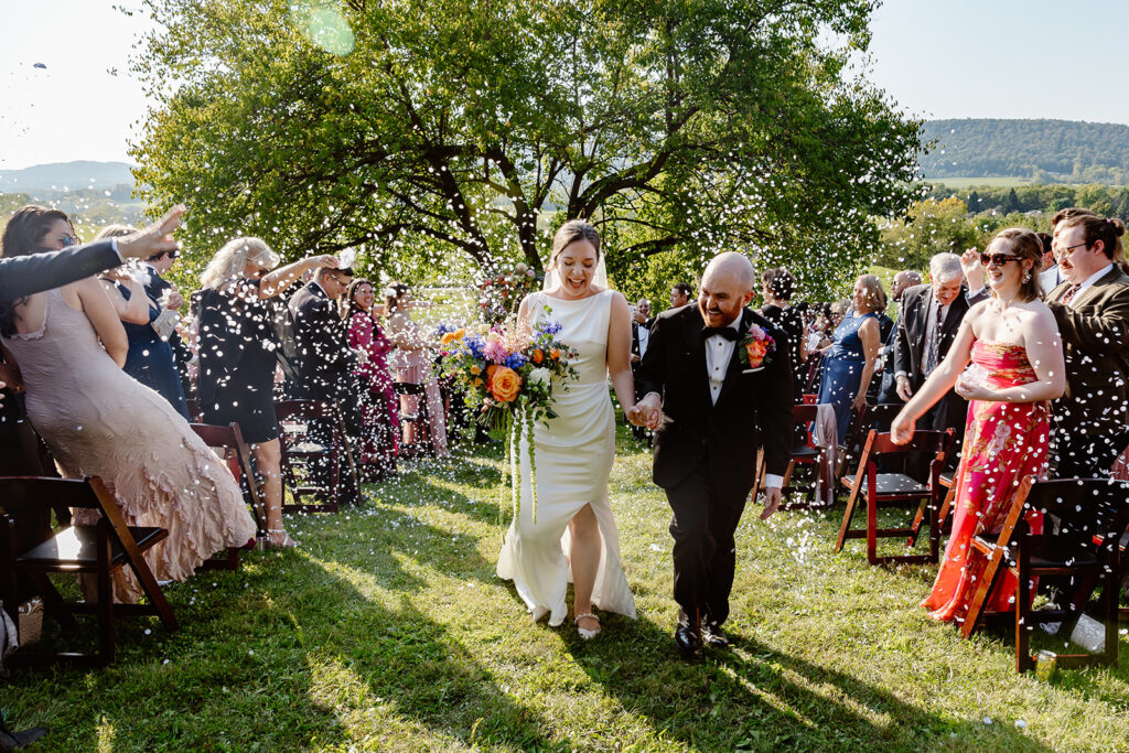 A bride and groom exiting their intimate outdoor ceremony. Guests are throwing biodegradable petals, and the couple is smiling. Behind them is a huge mulberry tree, and mountains. The bride is holding a large colorful bouquet, and wearing a sleeveless satin dress. The groom is wearing a Tux with a large, colorful boutonniere