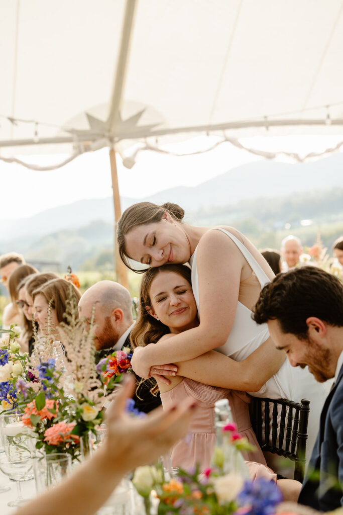 A bride leans in to hug her best friend during a wedding reception. The friend is sitting at her table. Both have their eyes closed with emotion. You can tell they are both so happy