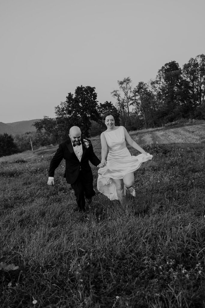 A black and white photograph, where a couple is running in a field. The bride is holding up her sleeveless satin dress while she runs. 