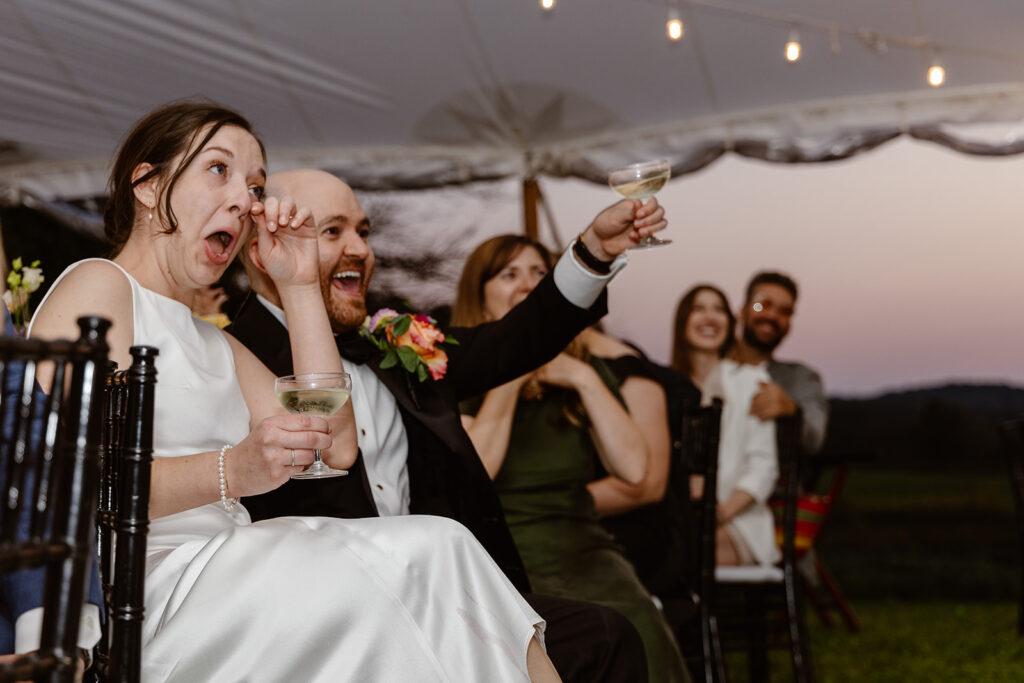 A bride and groom seated during a toast at their wedding reception. The bride is wiping tears away from her eyes. The groom is smiling an holding his glass up to toast. 