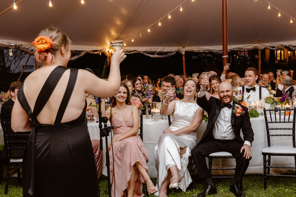 You can see the back of the person giving a toast, and the faces of the ride and groom and guests. The bride and groom are smiling, and holding up their glasses to toast