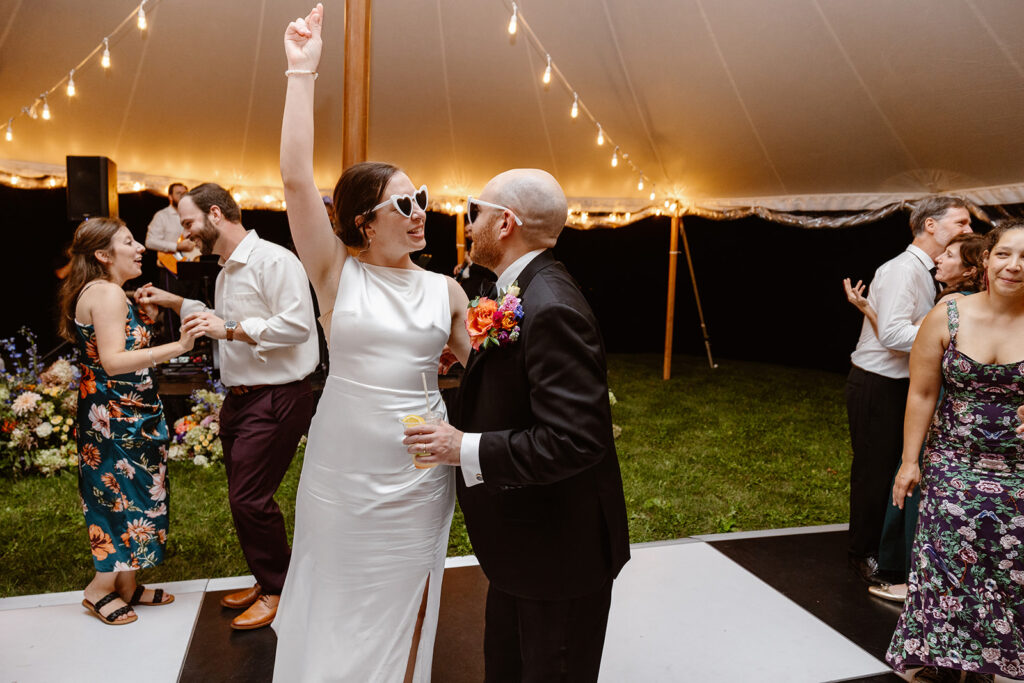 A bride and groom dancing during their reception. The bride and groom are both wearing heart shape sunglasses with white rims. the groom is holding a drink, and the bride hand is up snapping, They are on a black and white checkered dance floor, and the tent has twinkly lights.