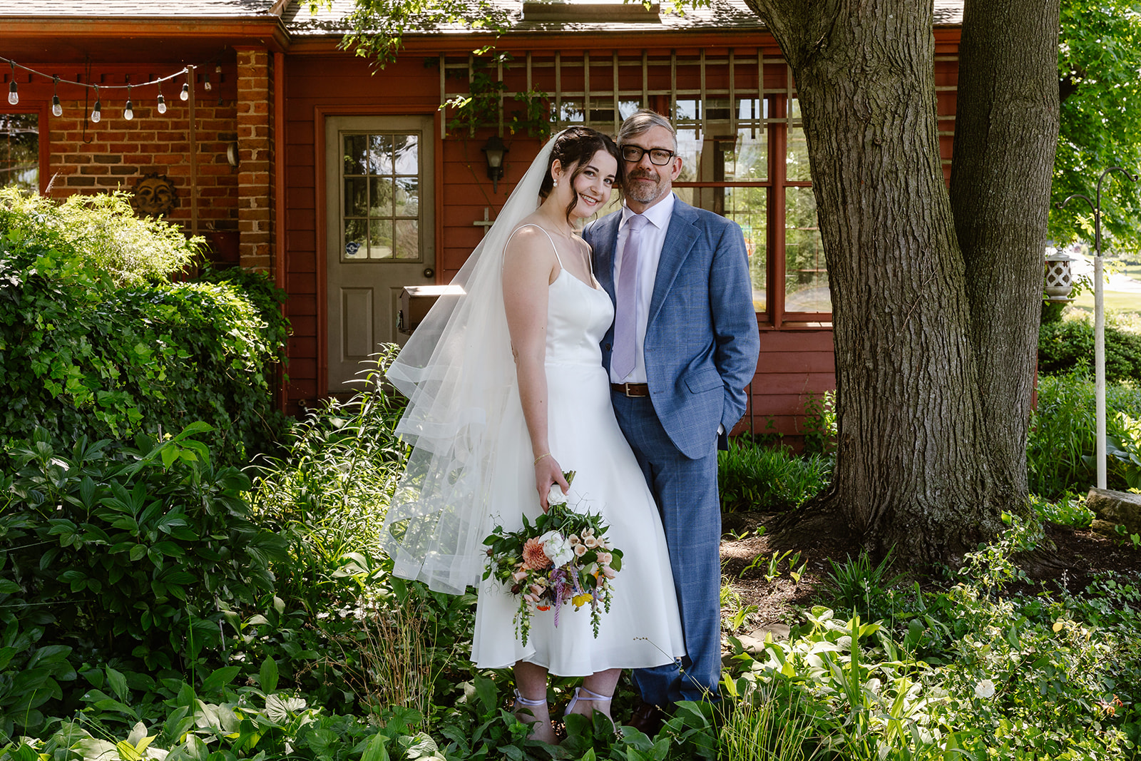 A couple standing in a yard the woman is wearing a white wedding dress and veil and is holding a bouquet of flowers and her partner is kissing her on the cheek and wearing a blue suit they are in a yard full of plants