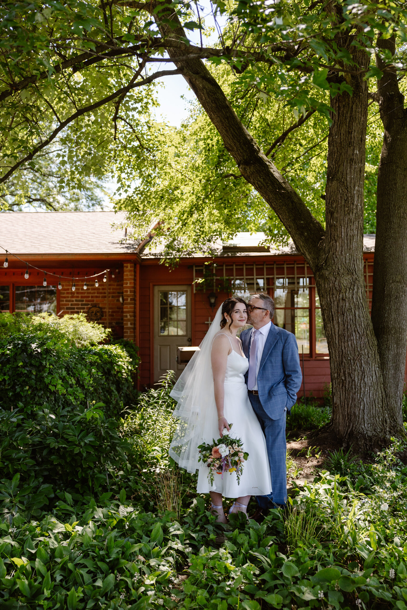 A couple standing in a yard the woman is wearing a white wedding dress and veil and is holding a bouquet of flowers and her partner is kissing her on the cheek and wearing a blue suit they are in a yard full of plants 