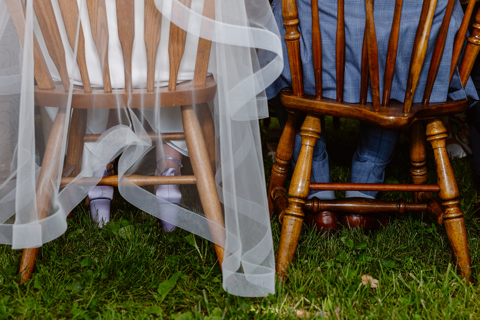 A closeup of a bride and groom sitting in mix matched chairs during their wedding reception. The brides veil is hanging over the chair, and you can see the details of her purple heels, and the grooms blue plaid suit.