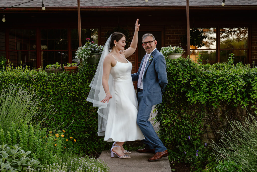 A bride and groom dancing during their portrait session during their backyard wedding. The bride is wearing a calf length satin dress with purple heels, and  a veil, and the groom is wearing a blue plaid suit with a purple tie