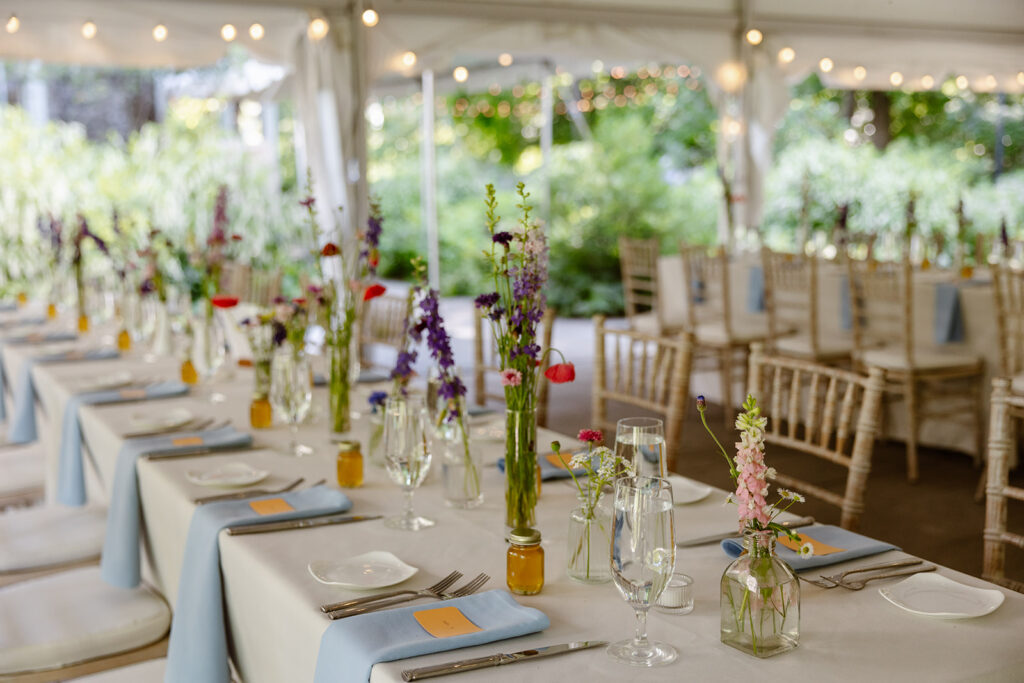 A set table at  a wedding reception. The table is in a tent. There are light blue napkins, and vases with poppies and colorful wildflowers. There are also jars of honey on the table