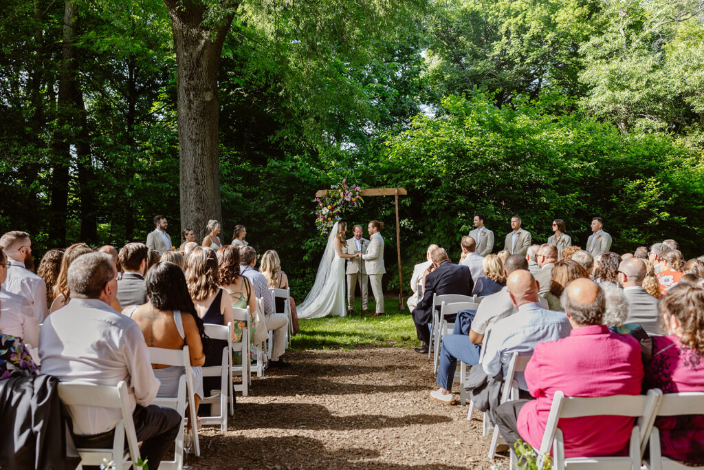 A bride and groom stand while holding hands during their intimate wedding ceremony. They are standing under an arbor that is decorated wioth gorgeous summer blooms