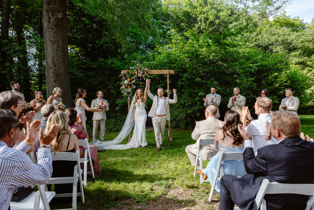 A bride and groom lift their hands in celebration after being announced bride and groom. The bride is wearing a Boho dress with a long custom veil, and the groom is wearing a tan linen suit. The ceremony is under an old tree