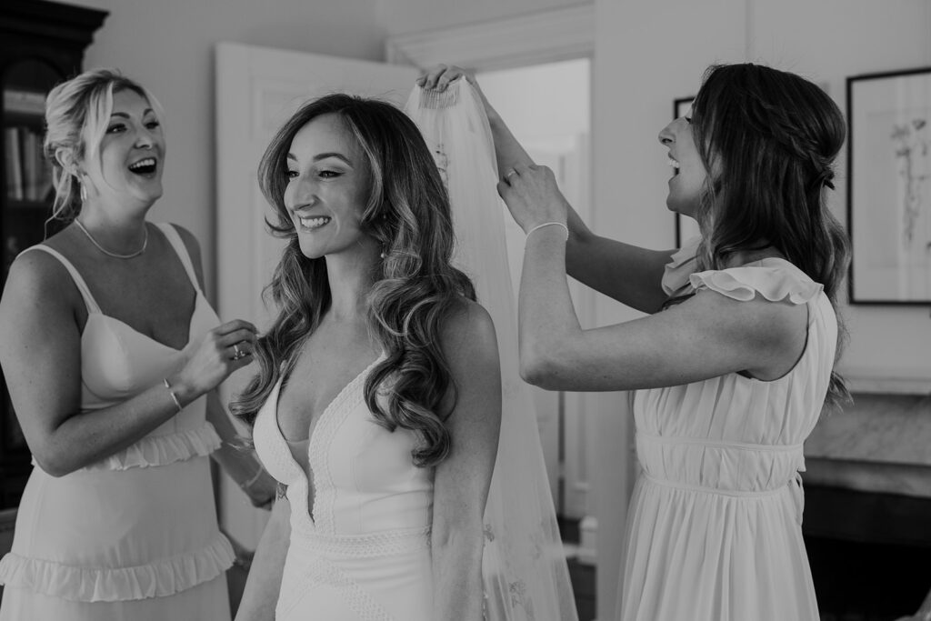 A black and white photo of bridesmaids helping the bride put on her veil. All three women have smiles on their faces