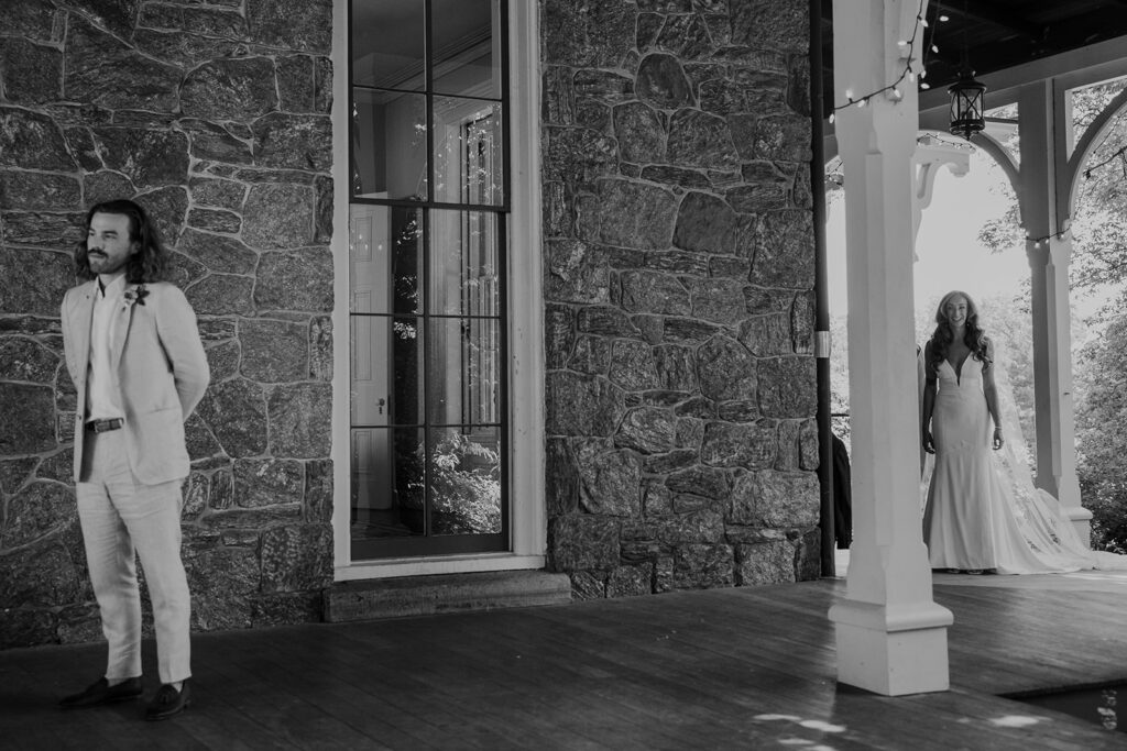 A black and white photo where the groom is waiting to see the bride for the first time on their wedding day. He looks nervous in the foreground, and bride looks excited as she is walking towards him.