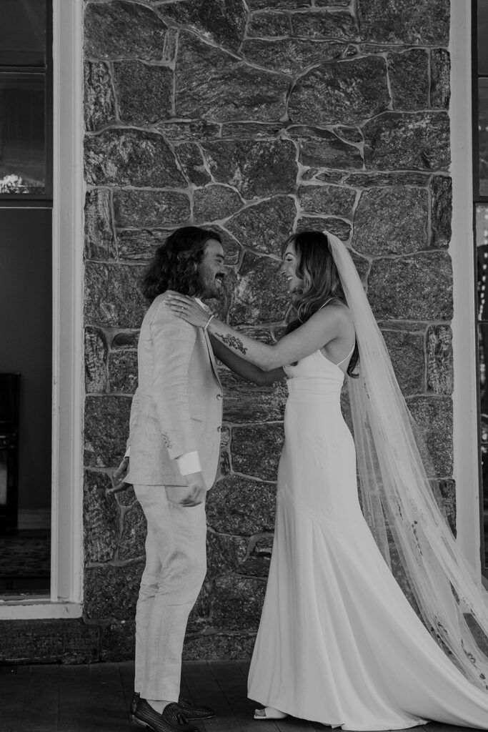 A black and whit photo of a bride and groom facing each other. The brides hands are on the grooms shoulders, and they are both beaming with joy