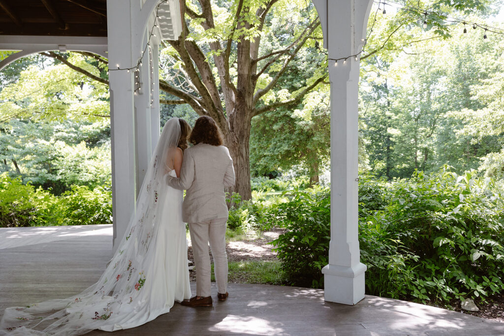 A bride and groom from behind during the reading of their private vows. The couple is standing on a victorian porch, and the groom has his hand gently resting on the back of the bride