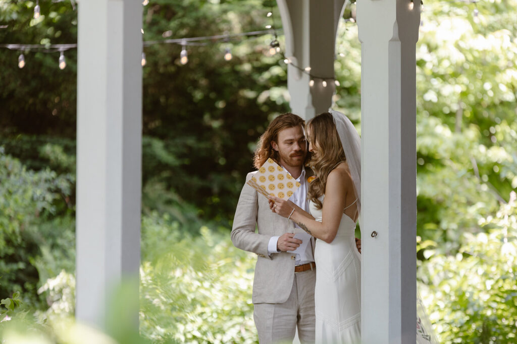 A bride is holding a booklet with her vows, and is reading them to the groom. He is looking at her lovingly as she reads. The groom is in a linen suit, and the bride is wearing a Boho dress