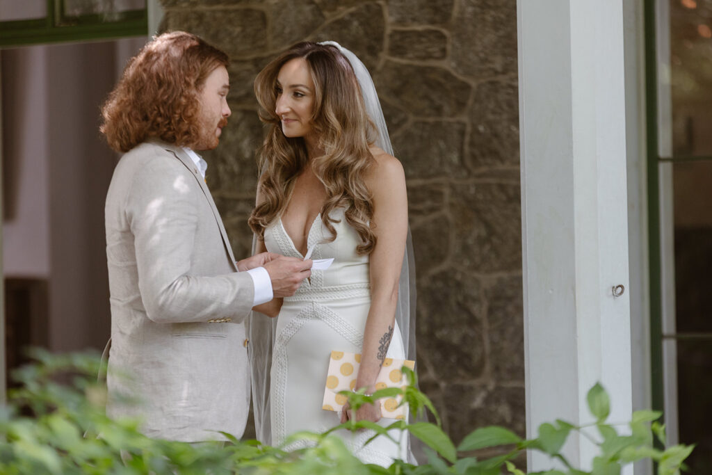 A bride listening to the groom read his personal vows. The couple is standing on the porch of a victorian mansion. 