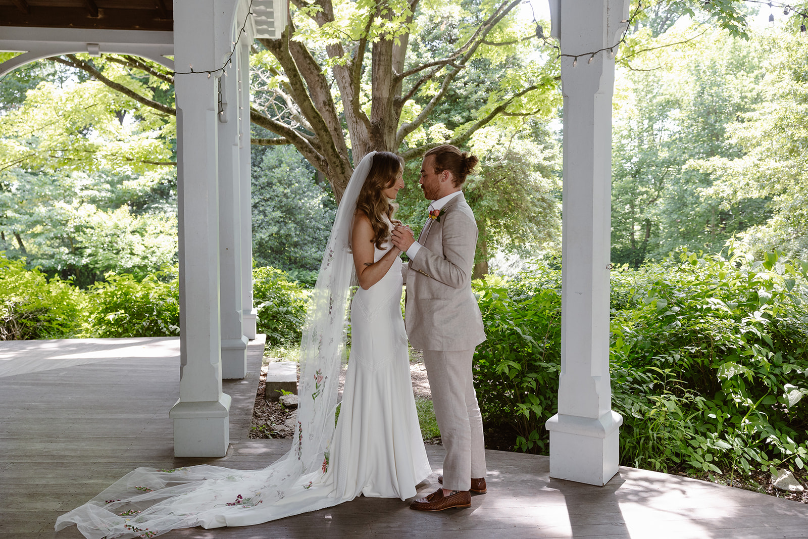 A bride and groom standing facing one another holding hands. The bride is wearing a Boho gown with an embroidered veil. The groom is wearing a tan linen suit. The couple is standing on an old Victorian porch in the outskirts of Philadelphia